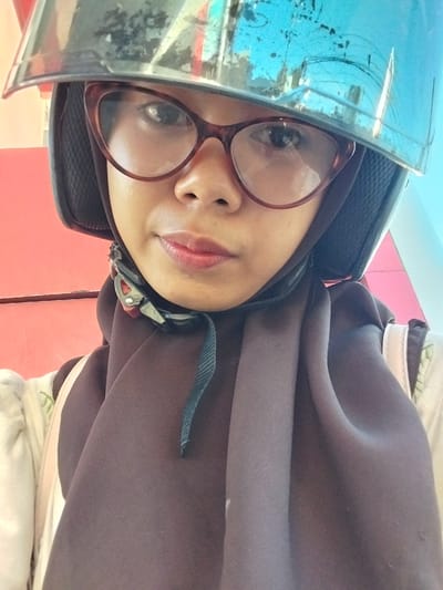 The media file is a selfie of a person wearing a helmet and glasses. The person is facing the camera and is partially obscured by a blue helmet with a reflective visor. They are wearing glasses with dark, thick frames. The person is also wearing a dark brown hijab or headscarf that covers their hair and neck. A portion of their face, including their eyes, nose, and lips, is visible. Their lips appear to be lightly colored with lipstick. The background of the image is blurred, but it appears to be an indoor setting with red and blue elements, possibly furniture or walls. The lighting suggests it could be daytime, but the specific time of day is not clear. The overall impression is that the person is either preparing for or has just completed a motorcycle ride, given the helmet. The image is taken from a close-up, low-angle perspective. There is no visible text in the media file.
