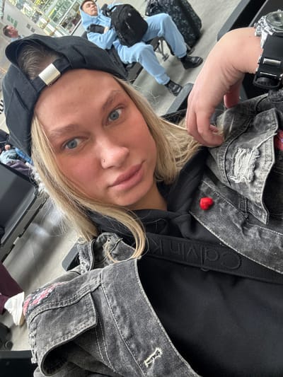 A selfie of a person with fair skin, blonde hair, and blue eyes, wearing a black hooded sweatshirt under a gray denim jacket with distressed details and a small red heart pin. They have a piercing on their nose. A black Calvin Klein-branded strap is visible across their chest. They are wearing a black digital watch. In the background, there is a person sitting with light blue clothes and luggage. The floor appears to be light gray tile. The location of Boldino, Russia is not apparent within the visible confines of this photograph.
