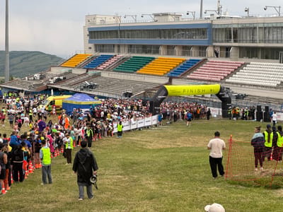 The image depicts a large outdoor gathering of people, likely a marathon or sporting event, at a stadium with tiered seating. The stadium has a modern facade with large windows and is backed by a hilly, vegetated landscape under an overcast sky. In the foreground, a grassy field is marked with orange cones and a red safety net. Several individuals are visible in the midground and background, including participants in athletic wear, volunteers in high-visibility vests, and spectators. A yellow inflatable archway with "FINISH" printed on it is prominently displayed, along with several event banners and tents. An ambulance is also visible, suggesting medical support for the event. The overall scene suggests an organized event with a substantial number of attendees, taking place in an outdoor stadium environment. The location is identified as Talgar, Kazakhstan.