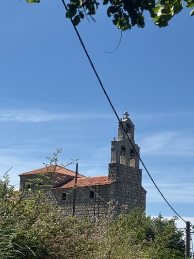The image shows a stone church with a red-tiled roof and a bell tower against a bright blue sky. The church appears to be located in a rural or semi-rural setting, with lush green foliage and trees in the foreground. A power line is visible stretching across the frame, partially obscuring the view of the church. The architecture of the church suggests it is an older structure. No people or animals are visible in the image.