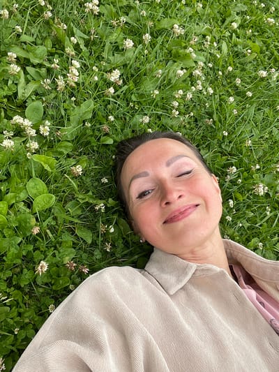 A woman lies on her back in a field of green grass and white clover flowers. She is wearing a light beige corduroy jacket over a pink top. Her left eye is closed in a wink, and she is smiling faintly. The image appears to be taken from a slightly elevated perspective, looking down on the woman. The location is Solov'evka, Russia, where the woman is enjoying a pleasant day outdoors.