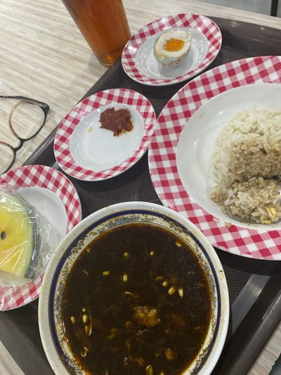 The image shows a tray of food on a wooden table, likely in a restaurant or food stall. The arrangement of food suggests a meal setting. In the foreground, a bowl with dark broth and visible ingredients like sprouts and possibly meat is present. To the right, a plate with white rice and a spoon taking a scoop is visible. A small dish containing a reddish paste, likely chili or sambal, is also on a checkered plate. Another small checkered plate holds a half of a salted egg. A piece of yellow fruit, possibly melon or papaya, wrapped in plastic, sits on a checkered plate to the left. A tall glass of amber-colored liquid, likely iced tea, is in the upper right corner. A pair of eyeglasses with brown frames rests on the table to the left of the food. The checkered plates have a red and white pattern. The overall setting appears to be indoors with neutral lighting. There are no people visible in the image. While the location is specified as Ngawi, Indonesia, there are no discernible landmarks or contextual clues within the image to confirm this specific location; it is a generic food setting.