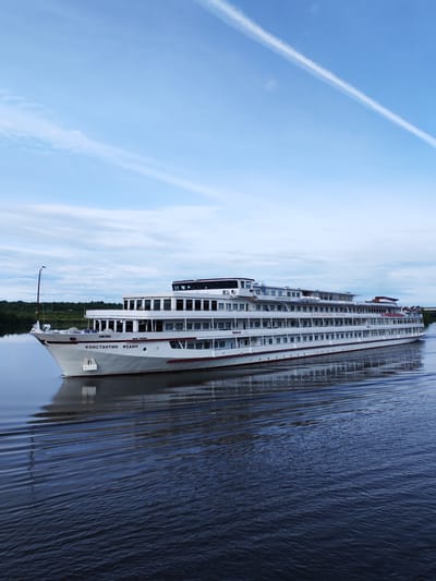 A wide, low-angle, full-shot photograph captures the multi-decked white river cruise ship "Konstantin Fedin" sailing on dark blue water, leaving a wake. The ship is positioned to the left of the frame, with its bow angled towards the viewer and its stern extending to the right. The vessel has multiple rows of windows along its sides, and several decks are visible, topped by a navigation bridge. Red trim accents the ship's hull and decks. In the background, a green, wooded shoreline is visible under a cloudy sky with a prominent white contrail. The location is identified as Krapivino, Russia.