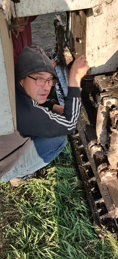 A man wearing a black hoodie with white stripes on the sleeves and blue jeans is lying on his back, appearing to work on a tracked vehicle. He is wearing glasses and looking towards the camera. The tracked vehicle is greasy and near some grass. The location is in Randava, Ukraine.
