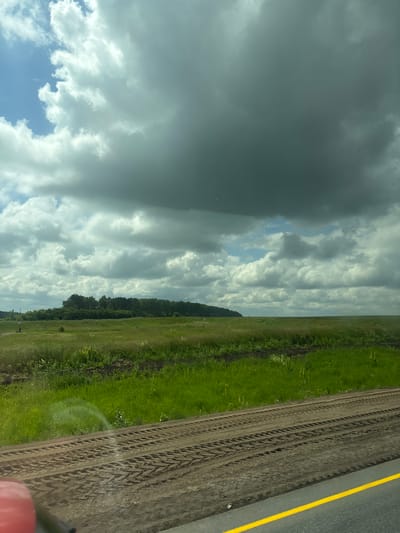 The image depicts a vast, open landscape under a dramatic sky. In the foreground, a dirt road with distinct tire tracks is visible, bordered by a dark gray asphalt road with a bright yellow line. To the left, a portion of a red object is partially visible, possibly part of a vehicle. Beyond the road, a field of tall green grass stretches towards a dense line of trees in the distance. The sky is filled with a mix of bright white and dark gray clouds, suggesting either a partly cloudy day with impending rain or the aftermath of a storm. The environment appears rural and undeveloped, typical of the outskirts of a small settlement. The location is identified as Таймурзино, Russia, which is a village in the Republic of Mordovia.