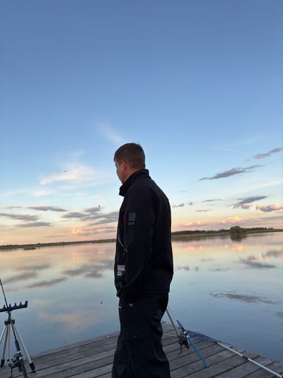 A medium, rotated, outdoor shot depicts a man in a black jacket standing on a wooden dock, near the shoreline of a body of water. The location may be Khryashchevka, Russia, based on potential visual cues like landscape or architecture.

The man is facing towards the water, with his head slightly bowed. Fishing rods are visible on the dock beside him, suggesting he is engaged in fishing.

The water reflects the sky, which features a mix of blue and cloudy patches. The time appears to be near dusk or dawn. The scene is static with no apparent interactions.