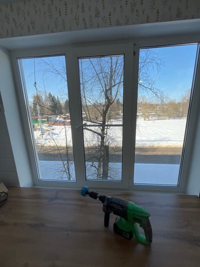 The image depicts an interior scene looking out through a large, white PVC triple-pane window. The window is set into a sloped wall with white trim, suggesting an attic or dormer room in Нарынка, Russia. Above the window, the wall is covered with wallpaper featuring a subtle pattern of light-colored botanical motifs.
In the immediate foreground, on a light brown wood-grain surface functioning as a deep windowsill, rests a green and black cordless hammer drill. The drill is positioned with its handle towards the right and its chuck, fitted with a blue circular brush attachment, pointing towards the left.
Beyond the window, a winter landscape is visible under a clear, bright blue sky. The ground is extensively covered in snow. A prominent bare deciduous tree with dark, intricate branches stands centrally in the view. To the left of the tree, in the middle distance, there are several small buildings with colored roofs (predominantly green and red) interspersed with bare trees and a cluster of evergreen trees. To the right, a broad expanse of snow-covered ground extends to the horizon, with a dark horizontal line traversing the middle ground, potentially a cleared road or dark area of ground. No individuals are visible in the scene.