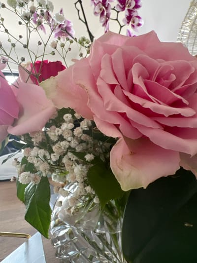 A close-up shot shows a bouquet of pink roses and white baby's breath flowers in a clear, faceted glass vase. The flowers are arranged in a way that the large pink rose is the most prominent subject, with its petals unfurling towards the viewer. In the background, blurred purple orchids are visible. The setting appears to be indoors, possibly on a wooden surface, with soft lighting. There are no people or animals present in the image. The image does not provide any information that can be used to determine its location in Bunbeg, Ireland.