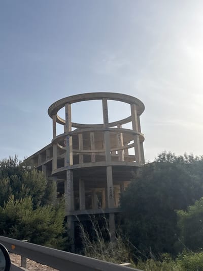 An unfinished, multi-story concrete building with a circular upper structure is surrounded by dense green foliage. The building's columns and beams are visible, and some of the concrete surfaces are marked with graffiti. The scene appears to be in an outdoor, possibly rural or semi-urban setting, with a clear sky and sunlight filtering through. A guardrail and part of a vehicle are visible in the foreground, suggesting the image was taken from a road. The location is identified as Urbanización La Cenia, Spain.