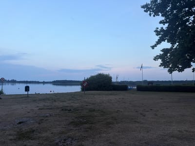 A tranquil, wide shot captures a lake at dusk in Preetz, Germany. The sky is a soft gradient of pale blue transitioning to lighter hues near the horizon, with faint wisps of clouds. The lake's surface is calm and reflective, mirroring the serene sky. In the foreground, a dry, mowed lawn stretches towards the water's edge.

Along the shoreline, several objects are visible: a red and white lifebuoy on a pole stands out, accompanied by a trash receptacle. Orange and white buoys mark a perimeter in the water, indicating a designated swimming or boating area. Beyond the immediate shoreline, a line of manicured green bushes forms a border. In the distance, sailboats with masts are docked or moored, suggesting a recreational area.

To the right of the frame, a large, dark tree with dense foliage partially obscures the sky. A flagpole with a German flag is also present in the mid-ground, adding a touch of national identity to the scene. The overall atmosphere is peaceful and quiet, characteristic of early evening in a suburban or recreational lakeside setting. No people are visible in the frame.