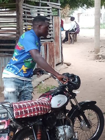 In Biliri, Nigeria, an individual stands next to a black motorcycle, holding onto the handlebars. The motorcycle features a white fuel tank with a "NIKE" logo. The seat is covered with a red and white patterned fabric. The person wears a blue patterned shirt and faded jeans. In the background, another person is seated on a stool-like bench and a weathered wooden structure is visible, as well as a dirt ground area.