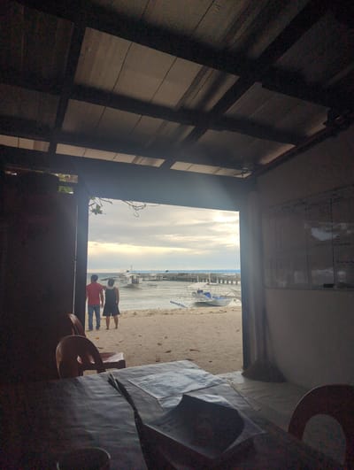 The image shows a view from inside a dark room or structure, looking out towards a beach in Logon, Philippines. Two people stand on the sand, one wearing a red shirt and the other a dark shirt. Several boats are beached on the sand, and a pier or jetty extends into the water. The sky is bright and cloudy. Inside the room, there is wooden paneling and a white board with writing on it. Part of a table and chair are visible in the foreground.