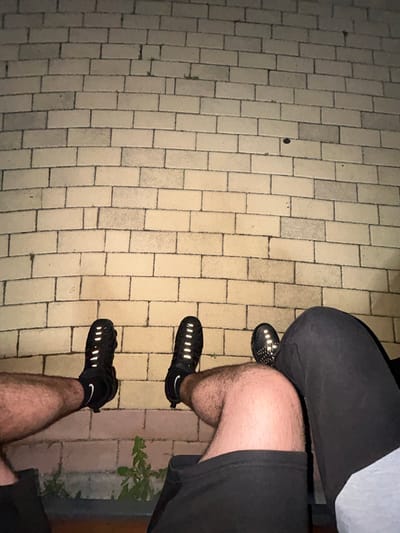 The image depicts a high-angle view of the lower bodies of two individuals sitting outdoors in Dzhebel, Bulgaria, at night. The foreground and background consist of a light-colored brick wall or paved surface with dark grout lines, illuminated by an unseen light source. Small patches of green vegetation are visible growing between some bricks.
The individual on the left has a visible hairy left leg, wearing what appears to be black shorts with a visible hem. The foot is shod in a black athletic shoe, specifically a Nike model with a visible white swoosh on a black sock, featuring multiple reflective horizontal strips across the top and side.
The individual on the right has a visible hairy right leg, wearing dark-colored shorts or sweatpants. The foot is also clad in a black athletic shoe with reflective horizontal strips, similar in appearance to the shoe on the left. A light blue or grey fabric garment, possibly trousers or sweatpants, is visible across this individual's lap, partially obscuring the upper thigh.
Both individuals appear to be seated with their legs extended forward, their feet positioned on or just above the brick surface. The reflective elements on the shoes are prominent, suggesting the use of a flash or direct light source during the photograph.