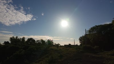 The image depicts a landscape under a bright, sunny sky, with the sun positioned high and slightly to the left of center. Wispy clouds are scattered across the blue expanse. In the foreground, there is a dark, grassy hill. Behind the hill, a dense line of dark green trees stretches across the middle ground, with some taller trees interspersed within. In the far distance, the silhouette of buildings and structures, including utility poles with wires, can be seen against the horizon. The overall impression is of an outdoor, possibly rural or semi-rural, setting bathed in natural sunlight. There are no people or animals visible in the frame. The location of Uruekanoe, Indonesia, is provided as context.