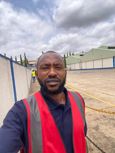 A man with a beard and a dark blue polo shirt and a red and gray reflective vest poses for a selfie. He is looking directly at the camera with a slight smile. Behind him, the scene is outdoors, with a paved area and buildings. The buildings appear to be industrial or commercial, with light green roofs and white walls. In the distance, other people can be seen, one of whom is wearing a bright yellow vest. The sky is cloudy. The ground is concrete with yellow and blue painted lines. There are some trees and a chain fence visible. The overall impression is that of a person on a work site. The image is taken from a low angle, looking up at the subject. The lighting suggests it is daytime.