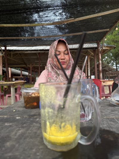 In Meunasahbale, Indonesia, a woman wearing a pink and green floral hijab and a pink top sits at a weathered wooden table outdoors. She is looking down at a small clear plastic container filled with a dark, chunky food item and possibly some red elements. In front of her, a large glass mug with a handle contains a yellow liquid with remnants on the side, and a black straw. Another glass, mostly out of focus, is visible to the right of the first. In the background, a rustic open-air structure with a corrugated roof and wooden supports is visible, suggesting a casual dining or seating area. The overall setting appears to be a food stall or outdoor restaurant, likely near a beach or coastal area, given the presence of a black mesh canopy overhead and vegetation in the distance.