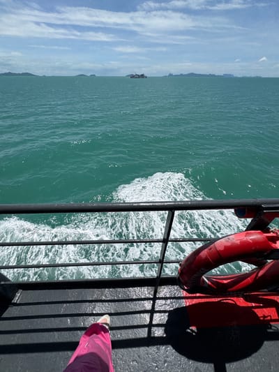 The image depicts a view from the deck of a ferry traveling on a bright, sunny day. The turquoise ocean stretches to the horizon, dotted with distant islands and a small cargo ship. The ferry is creating a white wake as it moves through the water. In the foreground, a black railing separates the deck from the sea, and a red life preserver is visible on the deck. A person's legs, wearing bright pink pants and white shoes, are visible at the bottom left of the frame, suggesting they are standing on the deck. The scene is indicative of travel by sea, likely a common mode of transportation for exploring the islands around Thong Nian, Thailand.