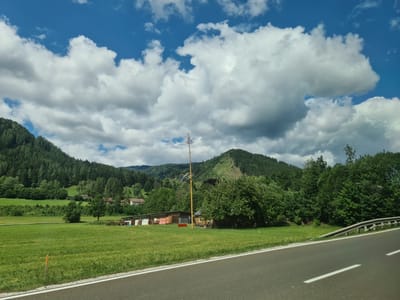 The image depicts a rural landscape with a road in the foreground, suggesting a drive-by perspective. A vast green field stretches across the middle ground, leading to a cluster of buildings and a tall wooden pole adorned with ribbons. The background is dominated by lush, tree-covered hills under a dramatic sky filled with white, fluffy clouds. The location is identified as St. Peter ob Judenburg, Austria, indicating a scenic, mountainous region. No people are visible in the frame. The scene appears to be a pastoral setting, possibly showcasing a traditional custom associated with the pole, which resembles a Maypole.