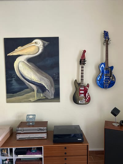The image depicts an indoor scene with a wall adorned with two electric guitars and a painting of a pelican. The pelican painting, rendered in a realistic style, shows the bird standing on a sandy bank beside a body of water under a dark sky. To the right of the painting, two electric guitars are mounted on the wall. The guitar on the left is black and red with silver hardware, while the guitar on the right is a vibrant blue with a mosaic-like pattern and silver hardware.
Below the guitars, on a wooden dresser or cabinet, several electronic components are visible. This includes a stack of what appear to be audio receivers or amplifiers, with one unit featuring a stand holding a digital device. To the right of these components, a black turntable is placed on the surface of the dresser. The dresser itself is made of wood and has several drawers.
In the lower left corner of the image, part of a pink book is visible, and below that, a white cube shelf unit contains various items, including books and what appears to be a small black box. On the far right side of the image, a portion of a wooden speaker cabinet is visible, with a small, cube-shaped object on a stand placed on top of it. The lighting suggests an indoor environment with ambient light. The overall scene suggests a room designed for listening to music or displaying musical instruments. The location is stated as Saint-Marcel-lès-Sauzet, France, though there are no explicit visual cues within the image to confirm this specific location.