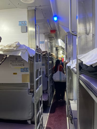 The image depicts the interior of a train carriage. The scene is characterized by rows of bunk beds on either side of a narrow aisle. Several bunk beds are visible, some with bedding made up, others appear empty. On the left side of the aisle, the beds are stacked three high, with metal railings for support. On the right side, the beds are also stacked, but the angle of the shot makes it difficult to determine the exact number of levels.

The aisle itself is carpeted with a dark red, speckled material. The walls of the carriage are a light color, possibly white or cream, and are adorned with various signs and labels. One prominent sign on the left wall features illustrations of two figures in a seating arrangement, with text that appears to be instructions. Another label with the number "51" is visible on the lower left. Above the beds, blue lights are visible, possibly indicator lights or part of the carriage's lighting system.

A woman is walking away from the camera down the aisle. She is wearing a dark top and dark pants, and is carrying a light-colored patterned bag over her shoulder. Her dark hair is pulled back. Further down the aisle, another individual, possibly male and wearing a blue garment, is partially visible in the distance. The overall impression is a functional, utilitarian interior of a passenger train, suggesting a journey in progress. There is no visible indication of the city of Sircaim, India, within the image itself.