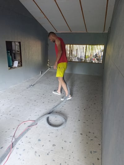 A man in a red tank top and yellow shorts is vacuuming a room with a terrazzo floor. The room has gray walls and a white ceiling with wooden beams. A window covered by vertical wooden slats is visible in the background, showing trees and people outside. There is a shelf with a blue gas cylinder and a white paper on the wall to the left. The vacuum cleaner has a gray hose and a red cord. The image appears to be taken indoors, possibly in an unfinished or newly constructed space.