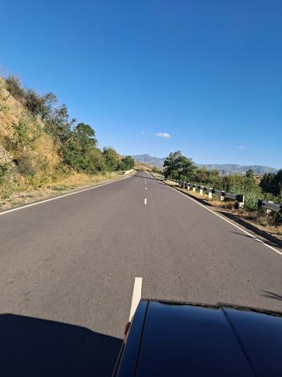The image shows a road in Getap, Armenia. The road is asphalt and has a white line down the center. On the left side of the road, there is a steep, grassy hillside with scattered trees and bushes. A metal guardrail with white reflective posts runs along the edge of the road, separating it from the hillside. In the distance, there are rolling hills and mountains under a clear blue sky with a few wispy clouds. The sun is bright, suggesting it is daytime, likely late morning or early afternoon, given the shadows. The perspective is from a vehicle driving on the road, as indicated by the partial view of a dark-colored car on the right side of the image. The car's surface is shiny, reflecting the surrounding scenery. The overall scene conveys a sense of travel and scenic beauty, with the road winding through a rural, mountainous landscape. There is no visible text in the image.