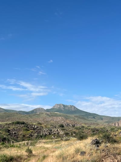 The image depicts a natural landscape under a bright blue sky with scattered white clouds. In the foreground, dry, golden grasses and scattered rocks suggest a semi-arid environment. A treeline with sparse bushes and darker green vegetation forms the middle ground, transitioning into a rocky, uneven terrain with more dense patches of greenery. Further in the background, rolling hills rise, culminating in a prominent, rugged mountain peak. The overall scene is one of natural, undeveloped terrain. While the location is stated as Malishka, Armenia, the image itself provides no specific urban or man-made features that would identify it as such.