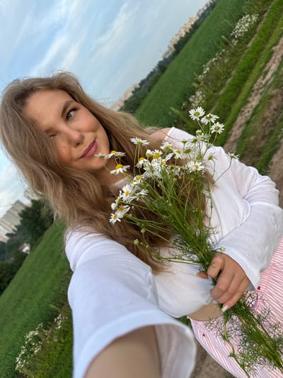 A young woman with long blonde hair is holding a bouquet of daisies. She is wearing a white long-sleeved shirt and a pink and white striped skirt. She is outdoors in a grassy area with some buildings in the background. The sky is cloudy. The location appears to be in the city of Сновицы, Russia.