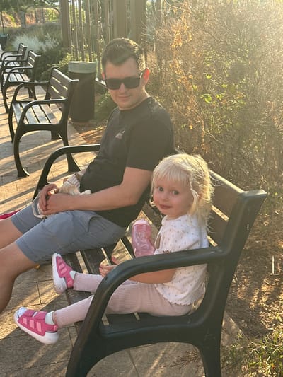 A man and a young child are sitting on a park bench outdoors. The man, wearing sunglasses and a black t-shirt, is looking towards the camera. The child, with blonde hair and a white patterned shirt, is also looking towards the camera, leaning forward. The child is wearing pink leggings and pink and white sneakers. A pink water bottle is visible near the child. The scene is bathed in the warm light of the late afternoon sun, casting long shadows. Trees and foliage are visible in the background. The setting appears to be a park with other benches in the foreground and a paved walkway. The atmosphere is relaxed and casual, suggesting a moment of rest during an outdoor activity. The location is Givat Shmuel, Israel.