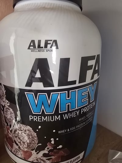The image shows a close-up of a white and black plastic container of "ALFA PREMIUM WHEY PROTEIN." The container is cylindrical and has large, bold lettering. "ALFA" is printed prominently in a large, black font on the left side, with "WELLNESS SPOR" printed below it in a smaller font. The word "WHEY" is printed in large, blue, outlined letters on the right side.
Below "WHEY," there is smaller text that reads "PREMIUM WHEY PROTEIN," and further down, "MIXES INSTANTLY" and "WHEY & EGG PROTEIN COMPLEX." On the far right, there is a strip of text with "IT FLAVOR" visible.
The background is a blurred image of what appears to be a wooden surface and some lighter gray areas. The lighting suggests an indoor setting. The product itself appears to be a supplement powder for fitness or wellness. There are no people or animals visible in the image. The container is tilted, giving a dynamic angle to the shot. The visual style of the label, with bold fonts and a contrasting color scheme, is typical of sports nutrition products.