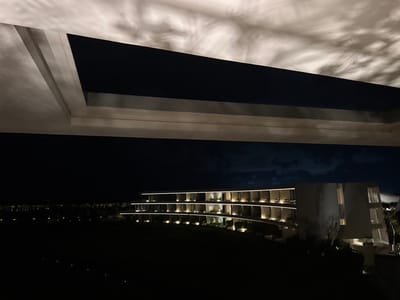 The image captures a modern building at night, seen from beneath a structure with intriguing light patterns. The building features illuminated balconies that curve gently. Below, a dark expanse suggests a lawn or field. Above, an overhang or roof detail shows shadows that resemble swirling clouds, creating a contrast between the geometric architecture and the soft, natural forms of the light play. The overall scene has a tranquil and mysterious ambiance.