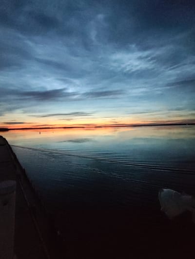 The image shows a calm body of water reflecting a twilight sky. The sky transitions from dark blue at the top to streaks of orange and pink near the horizon, indicating either sunrise or sunset. There are faint clouds scattered across the sky. The water is smooth, with a few subtle ripple patterns visible, suggesting a gentle disturbance.

Along the left side of the image, there is a dark, solid structure that appears to be a pier or embankment. Part of a white object, possibly a bollard or a light fixture, is visible on this structure. In the distance, two small, dark buoys are visible on the water.

The overall scene is serene and natural, with a focus on the interplay of light and water during the twilight hours. There are no visible people or specific landmarks that would definitively place the location within the city of Sheksna, Russia.