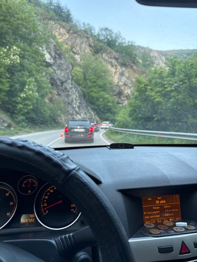 The image, taken from inside a moving vehicle, shows a winding road and mountainous landscape, likely in the vicinity of Бачковски манастир, Bulgaria, based on the dashboard display in Cyrillic script. The vehicle's dashboard is visible, displaying a range of 930 km, fuel consumption of 3.5 L/100km, and an instantaneous consumption of 0.6 L/h. The speedometer needle indicates a speed around 10 km/h, and the odometer reads 369722 km. Coins are placed on the dashboard. Through the windshield, one can see a vehicle ahead, marked by orange cones, suggesting road work or traffic control measures. The exterior landscape is dominated by a rocky hillside covered with green vegetation.