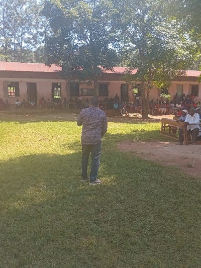 The image depicts an outdoor community gathering in Idakho South ward, Kenya. In the foreground, a male subject, viewed from the back, stands on a green grass lawn. He wears a dark, patterned long-sleeved shirt, dark blue jeans, and light-colored shoes. He faces a crowd of approximately 50-70 individuals, comprising men, women, and children, gathered in the midground. This crowd is seated on benches and directly on a reddish-brown dirt area in front of a single-story building. The building features light-colored walls, a reddish corrugated metal roof, and multiple open rectangular windows and doorways. Several large, leafy trees are positioned between the speaker and the building, partially obscuring parts of the assembled crowd and the structure. In the distant background, a dense stand of tall, slender trees is visible under a bright sky. The scene is illuminated by natural daylight, casting distinct shadows. The central figure appears to be addressing the assembled group.