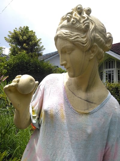 A weathered, cream-colored statue of a female figure is positioned outdoors in Ba Nwet Kone, Myanmar. The statue, depicted from the upper torso, gazes downwards and to its right at a spherical, light-colored object held in its left hand. Its sculpted hair is arranged in elaborate waves and curls. The statue's torso is covered by a real, light-colored, loosely fitting garment, possibly a t-shirt, which exhibits faint pastel pink, yellow, and blue dye stains. Lush green foliage, including tall grasses and shrubs, occupies the foreground around the statue. In the background, under a bright, clear sky, a white building with a dark, sloped roof is visible, featuring a prominent section of vertically segmented, multi-paned windows.