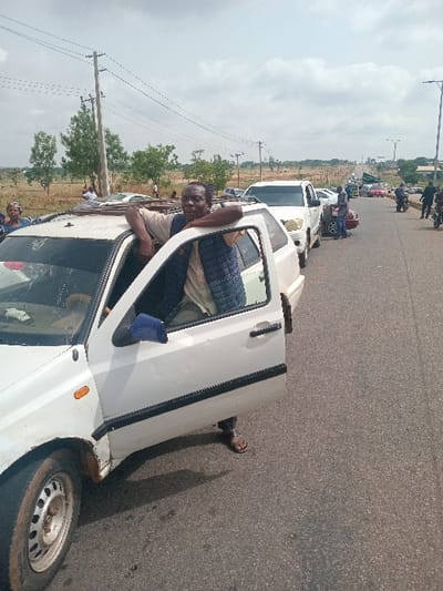 The image shows a man leaning out of a white Volkswagen Golf car. He is wearing a light blue shirt, a dark blue jacket, and dark sandals. The car's door is open and the man's feet are on the ground. The car is parked on a paved road with a line of other cars, including a white pickup truck. The man is looking at something off-camera. In the background, there are several other cars and people. There is a line of electric power poles along the side of the road. The scene is in Ungwan Tanko, Nigeria (10.422, 7.49) and there are trees lining the side of the road. The sky is overcast.