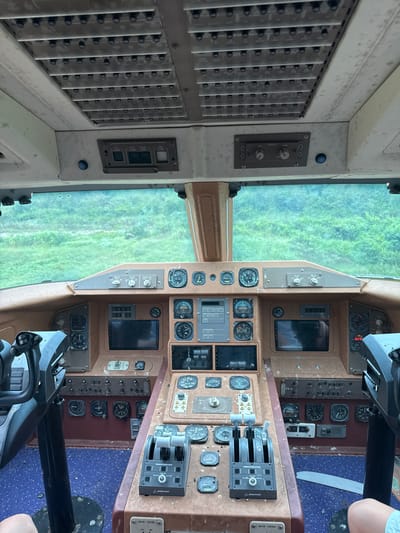 The image shows the interior of an old airplane cockpit, appearing to be a static display rather than a functional aircraft. The cockpit is furnished with numerous gauges, switches, levers, and two flight yokes. The instruments are dusty and worn, suggesting disuse. A blue carpet covers the floor of the cockpit. Through the windshield, a green, overgrown landscape is visible, likely a field or jungle, indicating an outdoor setting. The location is identified as Pak Nam Pran, Thailand. No people are clearly visible within the cockpit itself, although parts of limbs belonging to at least two individuals are present at the bottom edges of the frame, suggesting they are outside or partially inside the cockpit structure. The overall impression is of a decommissioned aircraft cockpit being utilized as an exhibit or attraction.