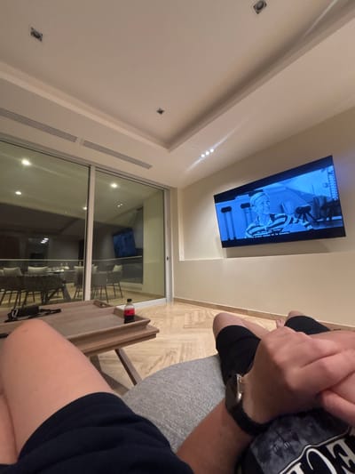 A person is sitting in a room with a TV mounted on the ceiling displaying a black and white image with Spanish subtitles. There is a sliding glass door leading to a patio with chairs and a table, suggesting a vacation rental or hotel room in Acapulco de Juárez, Mexico. The person has a watch on their left wrist. A small wooden table is located next to the chair. A bottle of Coca-Cola sits on the wooden table.