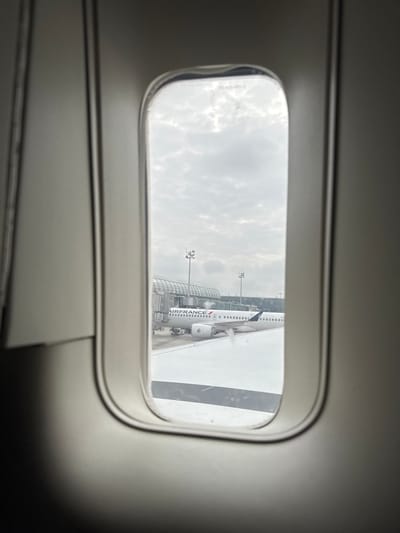A view from an airplane window shows an Air France airplane parked at Charles de Gaulle Airport in Le Mesnil-Amelot, France, under an overcast sky. Part of the wing of the airplane from which the photo is being taken is visible.