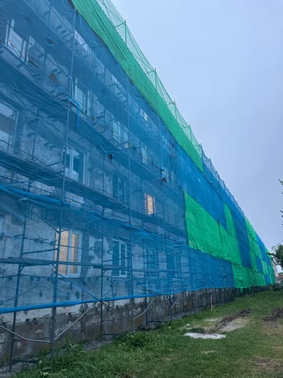 The image shows the exterior of a multi-story building undergoing renovation, covered by scaffolding and protective netting. The netting is a patchwork of blue and green, with visible scaffolding poles and platforms. Several windows are visible through the netting, some of which are illuminated from within. The building's facade appears weathered. The ground in front of the building is covered in grass, with some areas of disturbed soil. The sky is overcast. This scene is consistent with a construction or repair project in an urban or suburban setting.