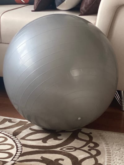 The image shows a large, grey exercise ball resting on a patterned rug in what appears to be a living room. The ball is positioned in the center of the frame and takes up a significant portion of the image. The rug has a cream-colored background with dark brown abstract patterns. In the background, part of a sofa or couch is visible, along with some pillows. To the right of the ball, a metallic leg of a piece of furniture is partially visible. The floor in the lower left corner appears to be made of wood. The scene is indoors, and the lighting suggests it's daytime. There are no people or animals in the image. The overall impression is one of a domestic setting. The location in Talas, Turkey, is an external detail that cannot be deduced solely from the visual information in the image.