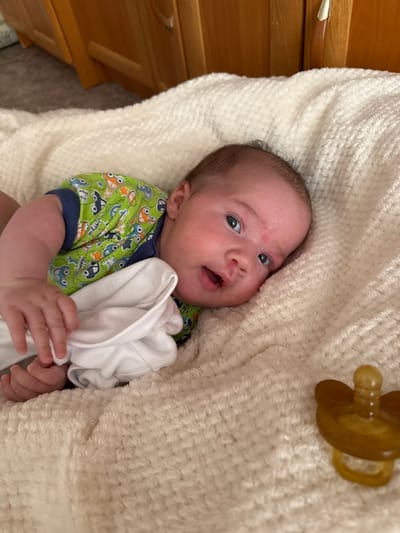 A newborn baby is lying on a cream-colored, fluffy blanket. The baby is wearing a green onesie with a pattern of cartoon cars and stars. The baby has dark hair and blue eyes and is looking upwards and to the right with its mouth slightly open. A reddish mark is visible on the baby's forehead. The baby's left hand is resting on a white cloth. In the lower right corner of the image, a brown pacifier is visible on the blanket. The background shows wooden furniture, likely a dresser or cabinet, in a warm tone. The scene appears to be indoors, possibly in a bedroom or living area.