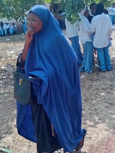 A group of people stand outdoors. In the foreground, a woman wearing a blue head covering and matching dress is visible. She holds something up to her mouth. A black bag hangs at her side. Behind her are other individuals, some wearing white shirts and blue pants. The ground is dirt and grass. Green foliage is visible on the left side of the frame. The image is oriented vertically.