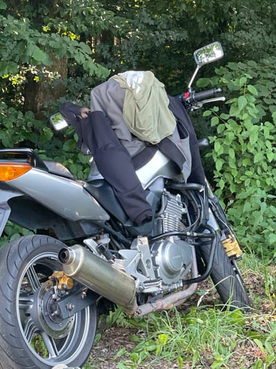 A silver motorcycle is parked on a dirt path in a wooded area. The motorcycle is partially covered with clothing, including a gray jacket and a light green t-shirt. The motorcycle's engine is visible, as is its exhaust pipe and rear wheel. The background consists of lush green trees and foliage. There are no people visible in the image. The scene is likely located in Beltershain, Germany, given the context provided.