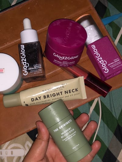 The image displays a flat lay composition of various skincare products on a brown, textured surface in an indoor setting in Tlogo, Indonesia. A light-skinned human hand, positioned in the lower right, holds a light green cylindrical "THE ORIGINOTE MUGWORT B3 CLAY STICK" (40g/1.41oz). Arranged on the brown surface are: a dark brown dropper bottle labeled "Glad2Glow AHA BHA PHA INTENSIVE PEELING SOLUTION" (15ml/0.5FL.OZ), a magenta jar of "Glad2Glow PEACH RETINOL MOISTURIZER" (30g/1.06FL.OZ), a magenta rectangular box of "Glad2Glow PEACH RETINOL SERUM" (17ml/0.57FL.OZ), and a beige tube of "NAAYA GREEN DAY BRIGHT NECK" cream. A red-tinted cylindrical tube, likely a lip product, is partially visible behind the moisturizer jar and neck cream tube. A white jar with a light blue cap is partially visible on the far left. The brown surface rests upon a fabric background featuring green, white, and dark blue vertical stripes.