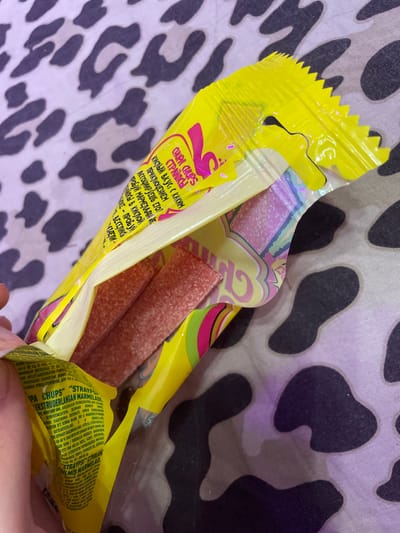 The image shows a close-up of an opened package of Chupa Chups "Straypsy" extruded fruit jellies, with the candies inside. The package is bright yellow and has a jagged opening. The candies appear to be red and covered in sugar. The background is a light-colored fabric with a black and gray leopard or cow print pattern. The scene is likely indoors and personal, given the nature of the product and the background. There is no visible information about the location in Borisova Griva Massiv, Russia.