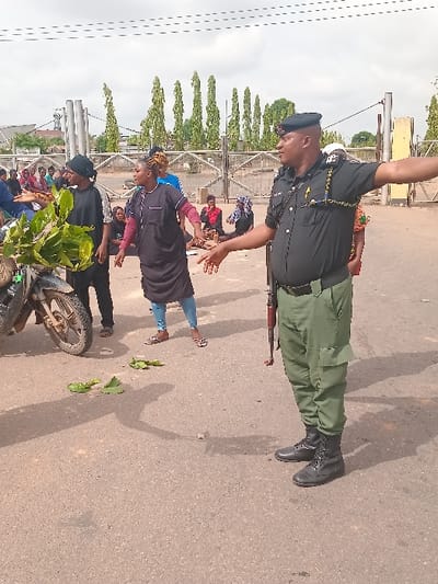 The image shows a scene in Ungwan Tanko, Nigeria, located at latitude 10.422 and longitude 7.49. A male officer, clad in a green uniform and black boots, stands with a rifle, facing a group of people. He is holding his right hand up in a gesturing manner, while his left hand rests on his waist.
The officer is positioned in front of a group of people, primarily women, some of whom are seated on the ground. A motorcycle, with a large leafy branch on the seat, sits on the left of the frame. Some leaves are scattered on the ground, and there are other objects, including a bag, visible in the background.
The scene appears to take place on a paved road, with a metallic gate and a row of trees behind it. The sky is partly cloudy with a visible white, cottony cloud formation.