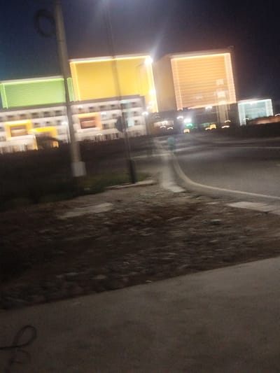 The image depicts an exterior night scene, likely a street or highway at dusk or night. In the background, a series of modern buildings are illuminated with colored lights. From left to right, the first building section is lit with green, followed by a large section in yellow, and then another building in a warm orange hue. To the far right, a smaller building is lit in a light blue. These structures appear to be hotels or similar large establishments, with visible rows of windows.
The foreground of the image shows a paved road or pathway curving into the distance. To the left of the road, there is a dark, unlit area that appears to be a grassy or unkempt embankment. A vertical pole, possibly a lamppost or utility pole, is visible on the left side of the image, with a circular object at its top. The overall lighting suggests it is nighttime, and the clarity of the image is low, indicating a potential long exposure or low light photography. There is no visible text in the image. The scene conveys a sense of urban or roadside development at night, with the illuminated buildings being the dominant feature. The location context provided, "Түндүк айланып өтүүчү жол 84 га, Kyrgyzstan," suggests this is a road in northern Kyrgyzstan.