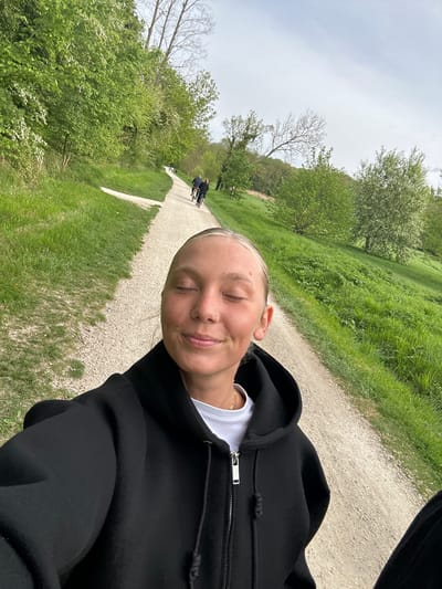 A woman takes a selfie on a gravel path lined with grass, trees, and other greenery on what appears to be an overcast day. She is wearing a black zipped hoodie with a white t-shirt underneath. In the background, down the path, two people are riding bicycles in Noisiel, France.