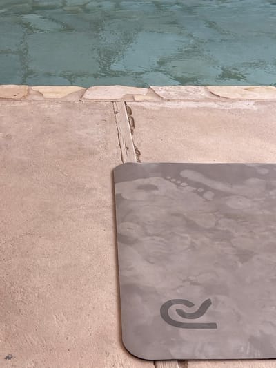 The image displays a scene at the edge of a body of water within the Arava Tichona Regional Council, Israel. The upper half of the frame shows a light blue-green body of water with a rippled surface. Immediately below, a border is formed by a row of irregular, light-toned stone or concrete flagstones. Beneath these flagstones, a textured light tan concrete or paved surface extends into the foreground. A narrow, weathered wooden slat is vertically embedded within a crevice in the concrete surface, situated just below the stone border. In the lower right quadrant of the image, a grey rectangular mat with rounded corners is positioned on the concrete pavement. The mat's surface exhibits numerous light-colored wet footprints and smudges, consistent with bare feet having recently contacted the mat after being wet. A dark grey, stylized 'R' logo is visible in the bottom right corner of the mat. No individuals are present in the scene.
